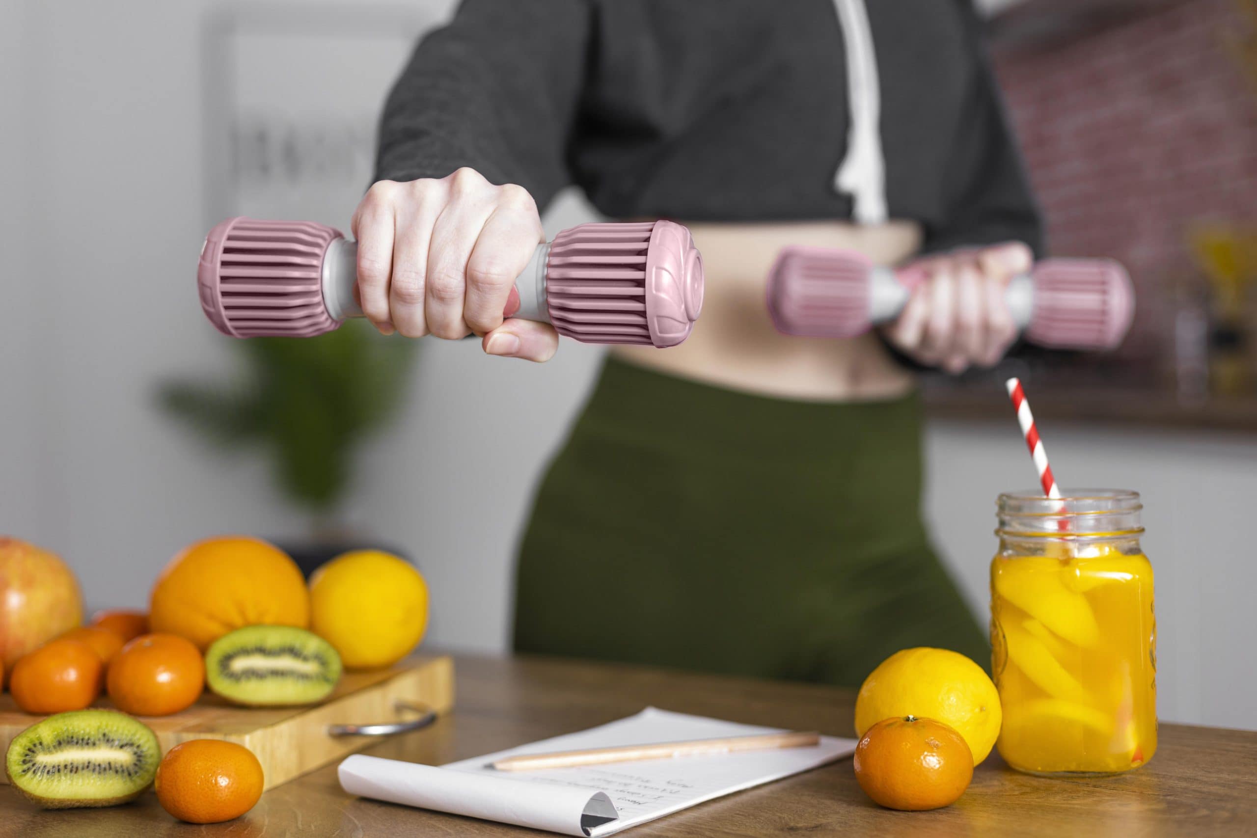 Close-up of a woman holding dumbbells with fresh fruits, juice, and a notepad on the table, illustrating the concept of Food as Medicine for Women through nutrition and exercise.