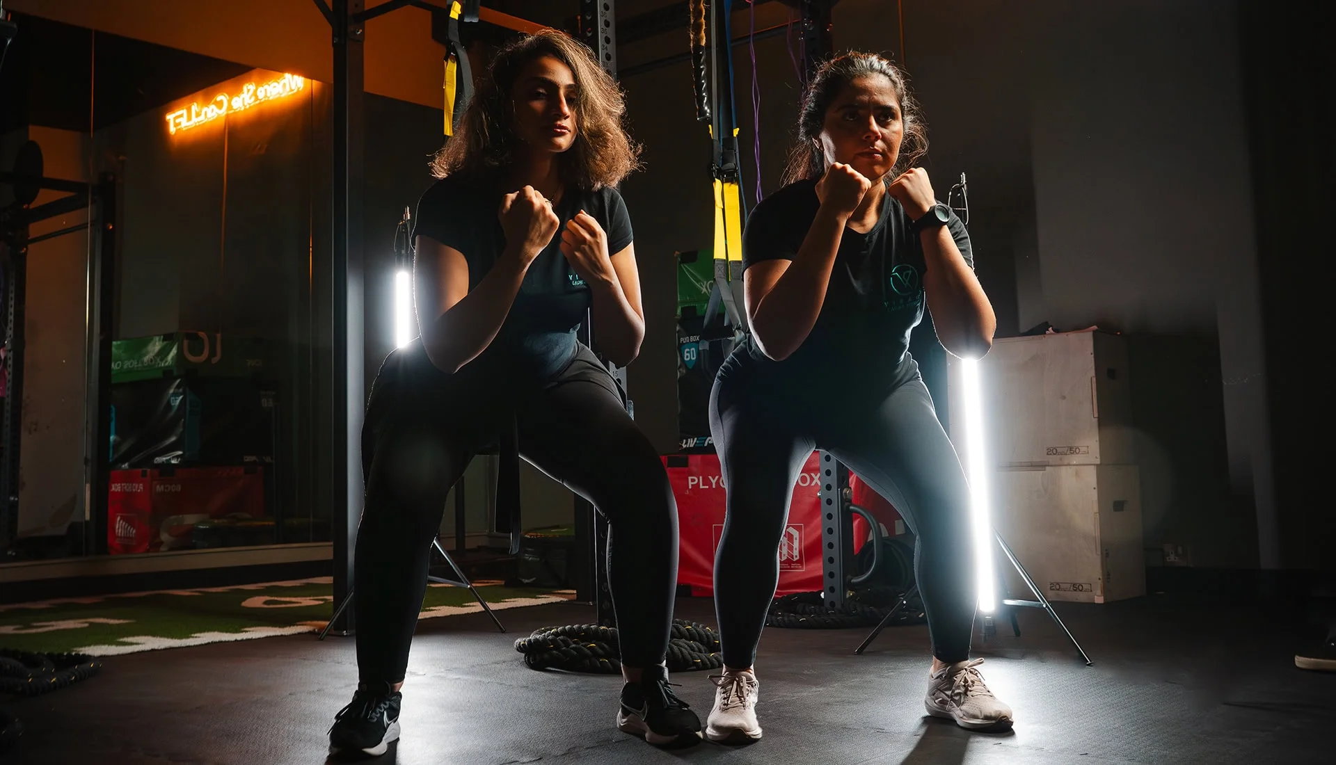 Two women in athletic wear performing synchronized squats in a dark gym, focused on strength and conditioning training